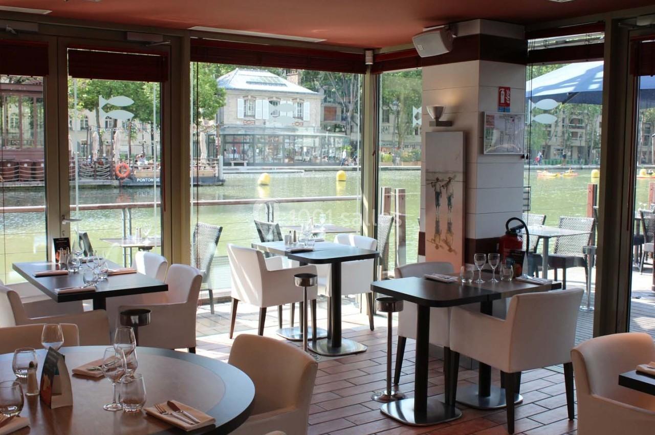 Salle de restaurant lumineuse avec vue sur un canal, tables dressées et grandes baies vitrées ouvertes.