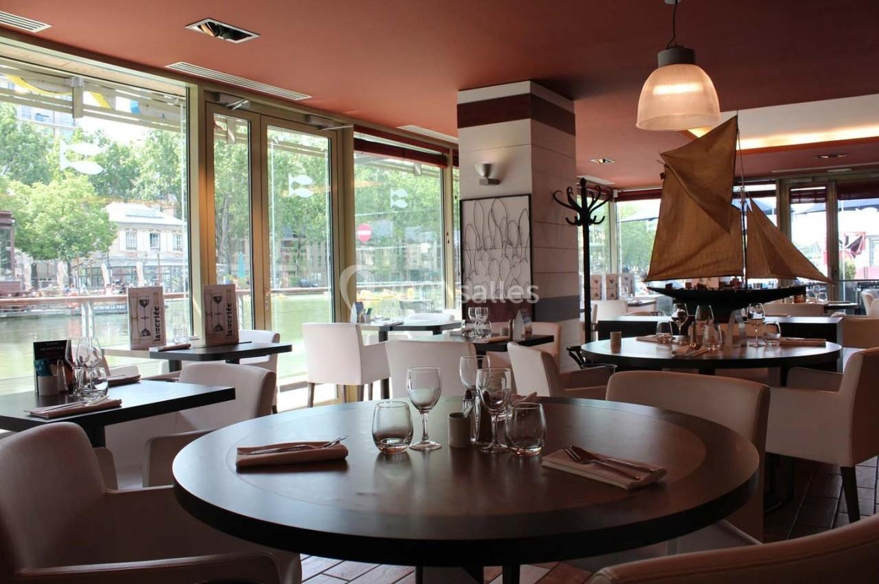 Salle de restaurant lumineuse avec grandes baies vitrées donnant sur un canal, tables dressées et décoration nautique.