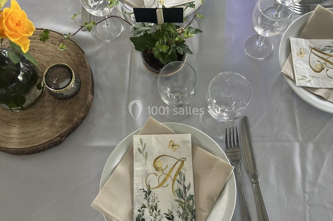 Table dressée avec vaisselle blanche, serviettes beiges, menus décorés de motifs floraux et centre de table végétal.