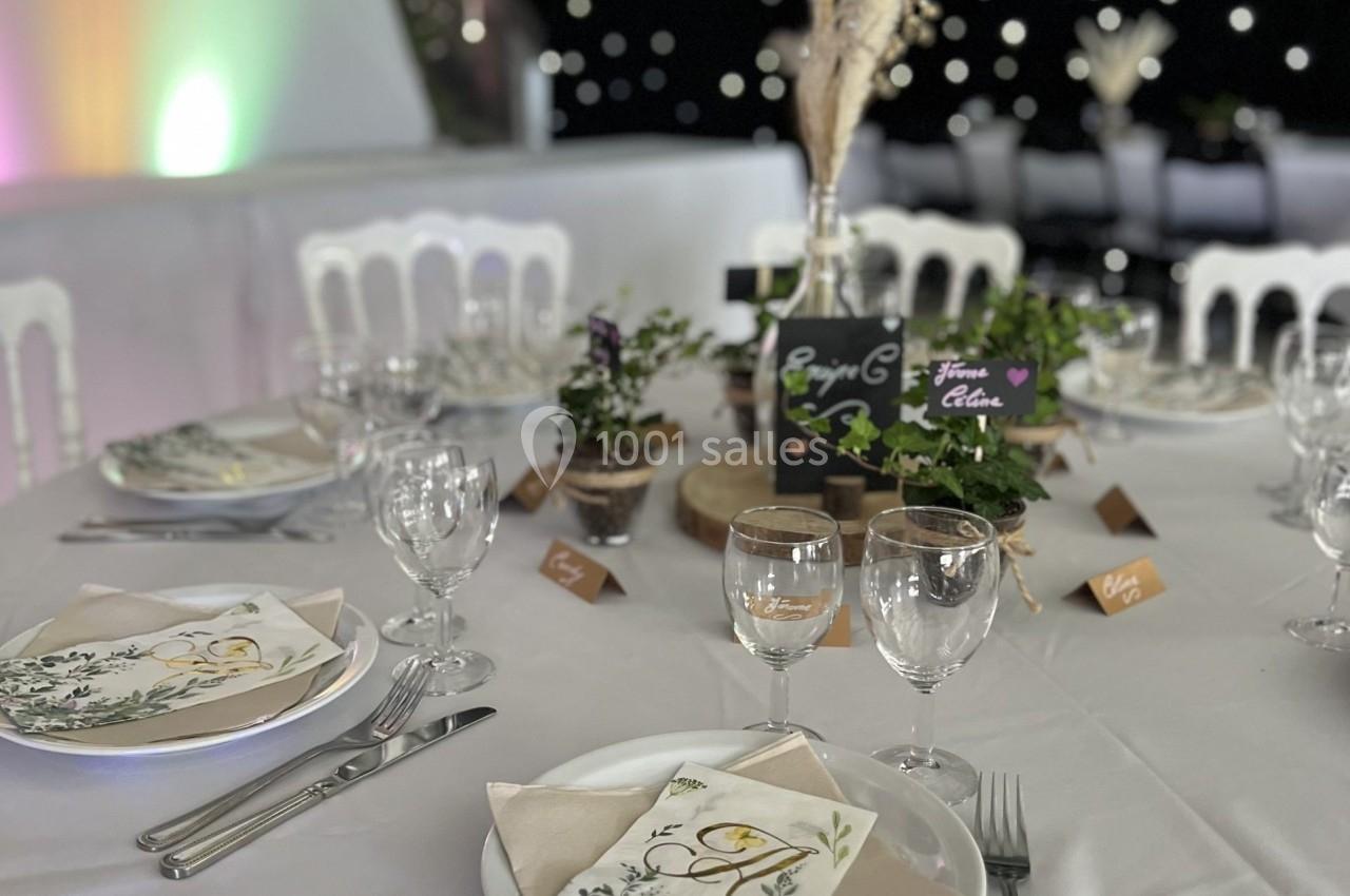 Table décorée pour un événement avec nappes blanches, menus illustrés, plantes et centre de table en bois.