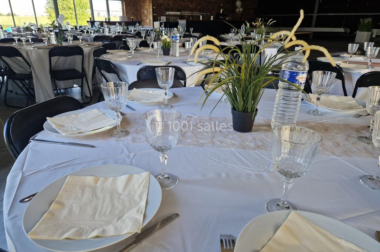 Tables rondes dressées avec nappes blanches, verres, assiettes, serviettes et centre de table végétal dans une salle…