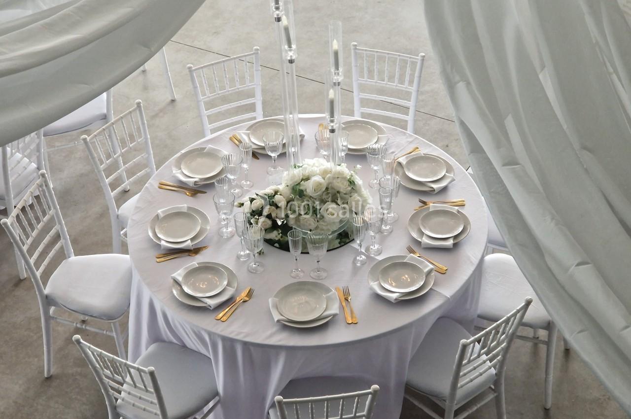 Table ronde élégamment dressée avec nappes blanches, chaises assorties, vaisselle raffinée et centre de table floral.