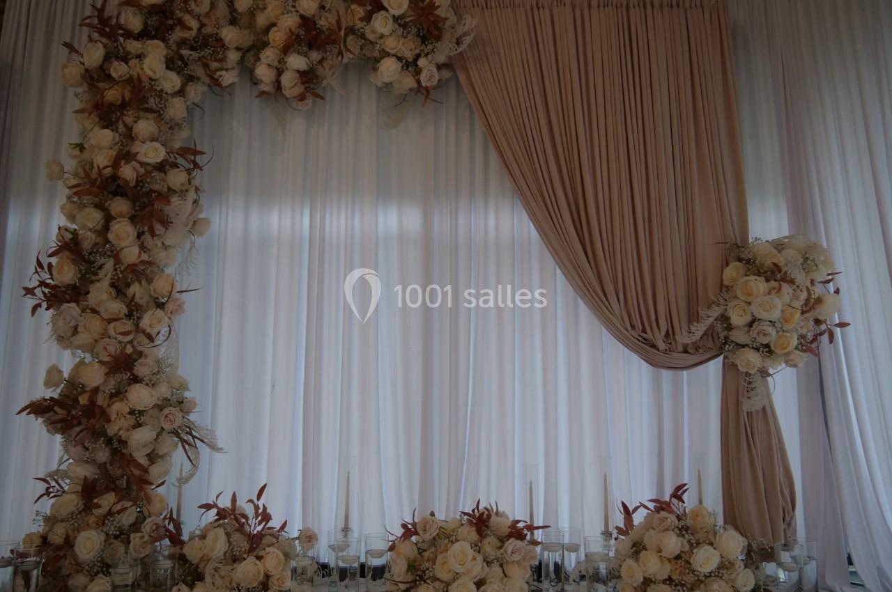 Décor floral avec des roses blanches et beiges formant une arche, accompagné de rideaux beiges et blancs en arrière-plan.