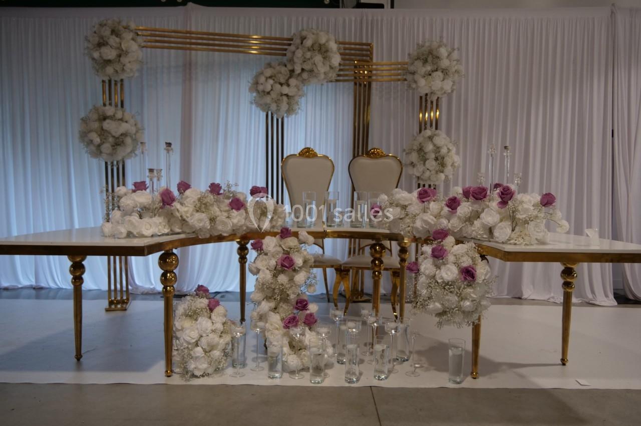 Décoration de mariage avec des chaises dorées, des tables ornées de fleurs blanches et violettes, et des vases en verre.