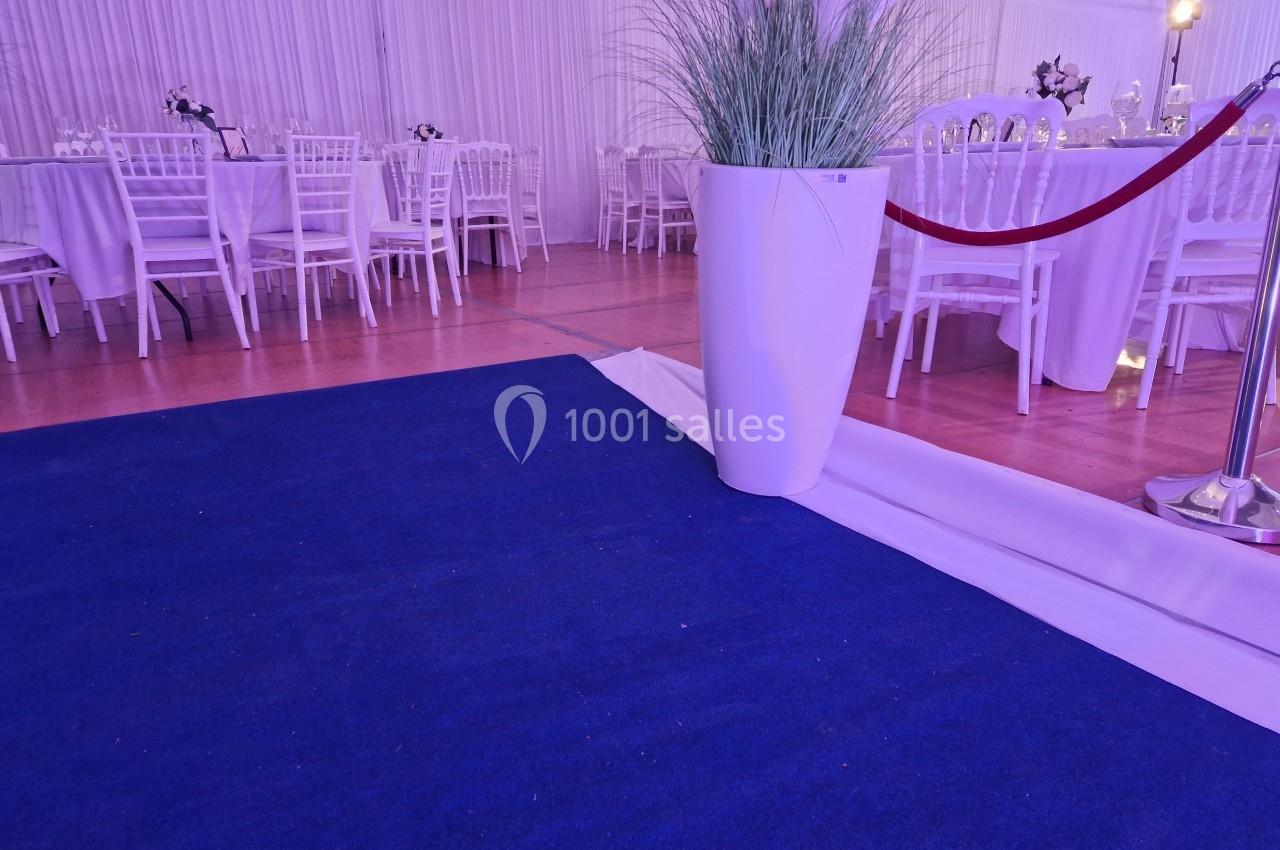 Tapis bleu menant à une salle de réception avec tables dressées et chaises blanches, décorée de plantes et d'une corde rouge.