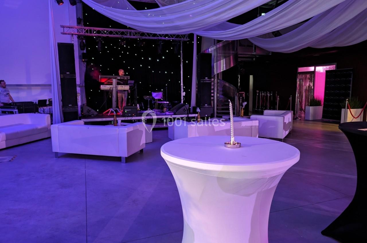 Salle événementielle avec scène éclairée, tables hautes blanches et canapés disposés autour.