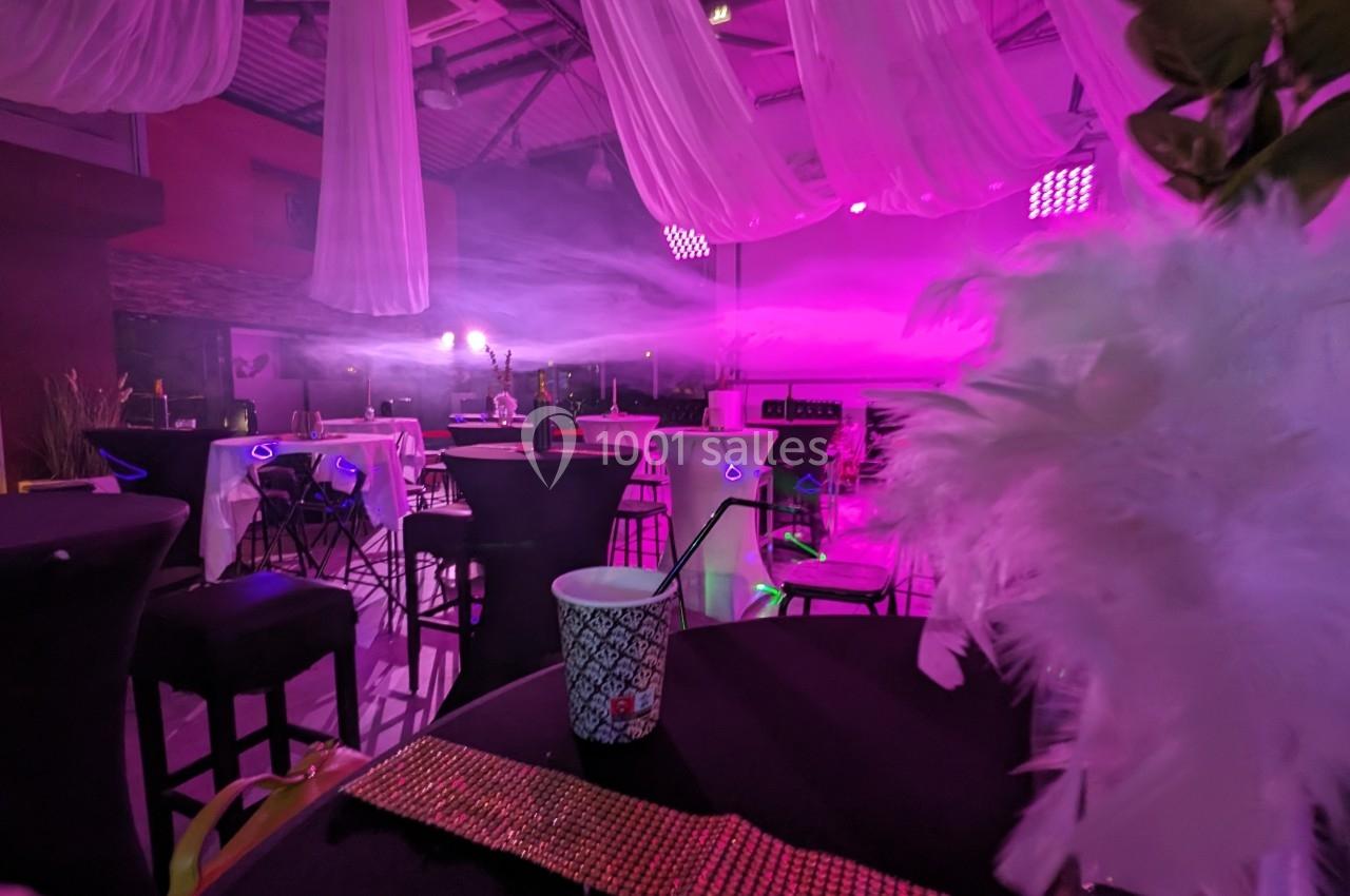 Salle décorée pour un événement avec éclairage rose, tables hautes, chaises noires et éléments de décoration blanche.