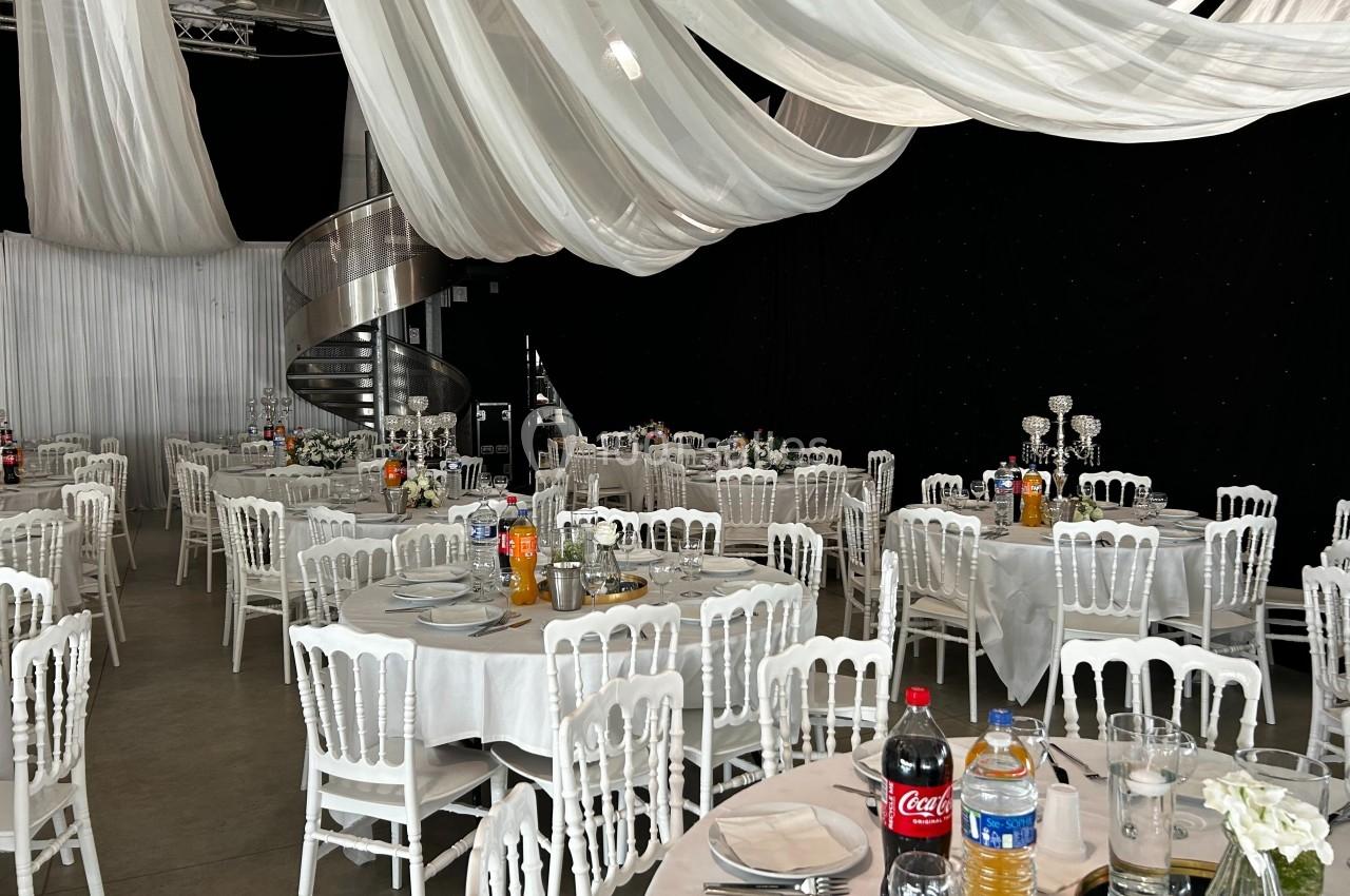 Salle de réception décorée avec des tables rondes dressées, chaises blanches et drapés suspendus au plafond.