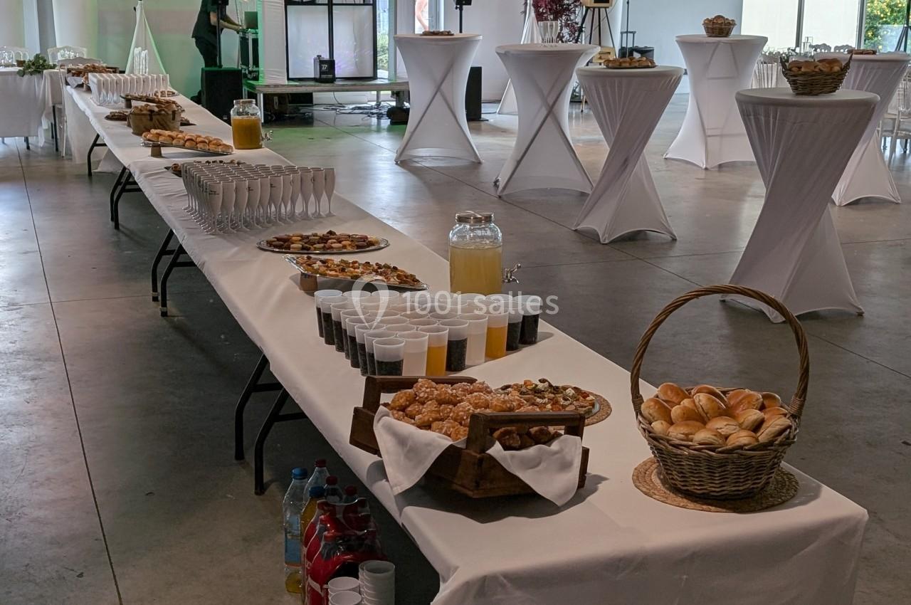 Table dressée pour un buffet avec viennoiseries, jus de fruits, verres et paniers de pain dans une salle lumineuse.