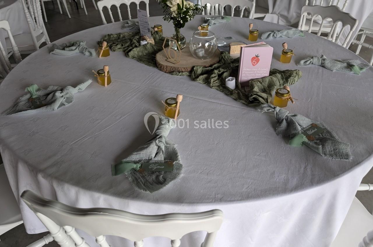Table ronde dressée avec une nappe blanche, décorée de petits pots de miel, serviettes vertes et un centre de table floral.