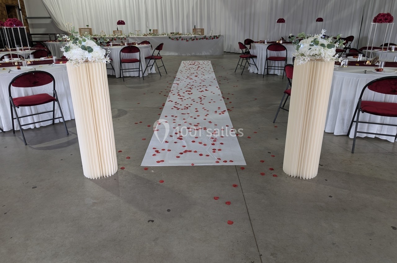 Allée centrale décorée de pétales rouges, encadrée de colonnes fleuries, menant à une table dans une salle de réception.