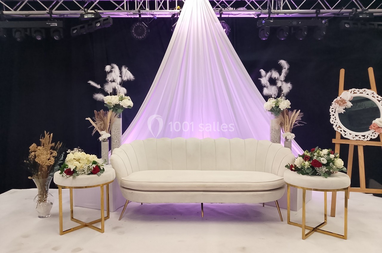 Canapé blanc entouré de tables décorées de fleurs, avec un fond drapé blanc et éclairage violet.