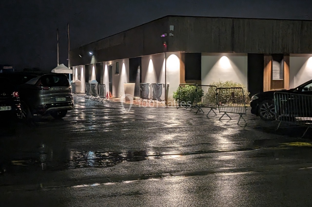 Parking humide de nuit devant un bâtiment éclairé, avec des barrières métalliques et des voitures stationnées.