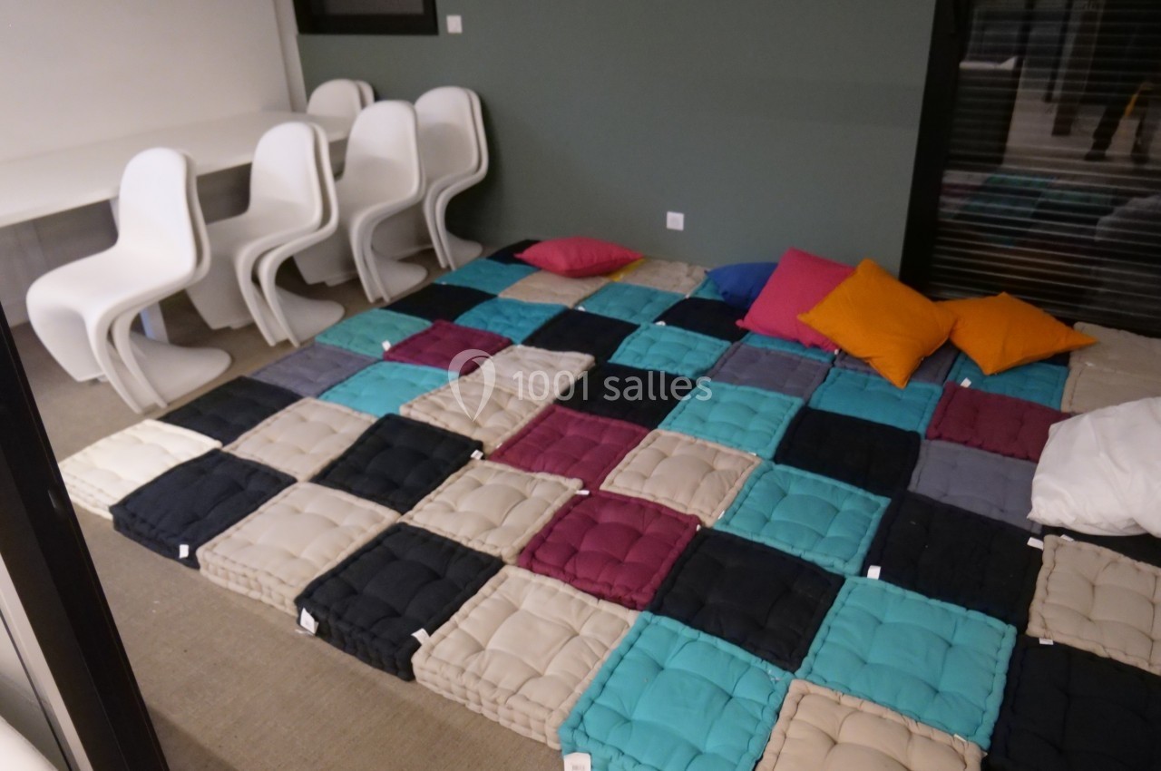 Coussins colorés disposés en damier sur le sol, entourés de chaises blanches et d'une table dans une pièce moderne.