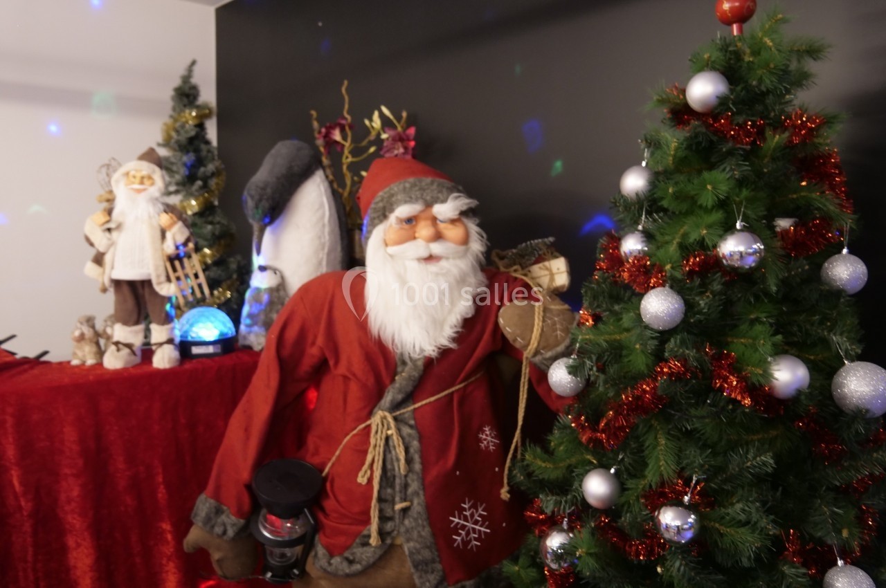 Un Père Noël décoratif près d'un sapin orné de boules argentées et de guirlandes rouges, dans un décor festif.