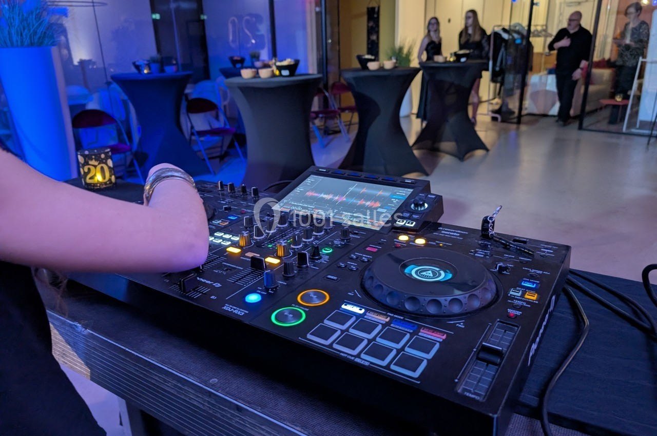 Un DJ mixe de la musique sur une table de mixage dans un espace moderne avec des invités en arrière-plan.