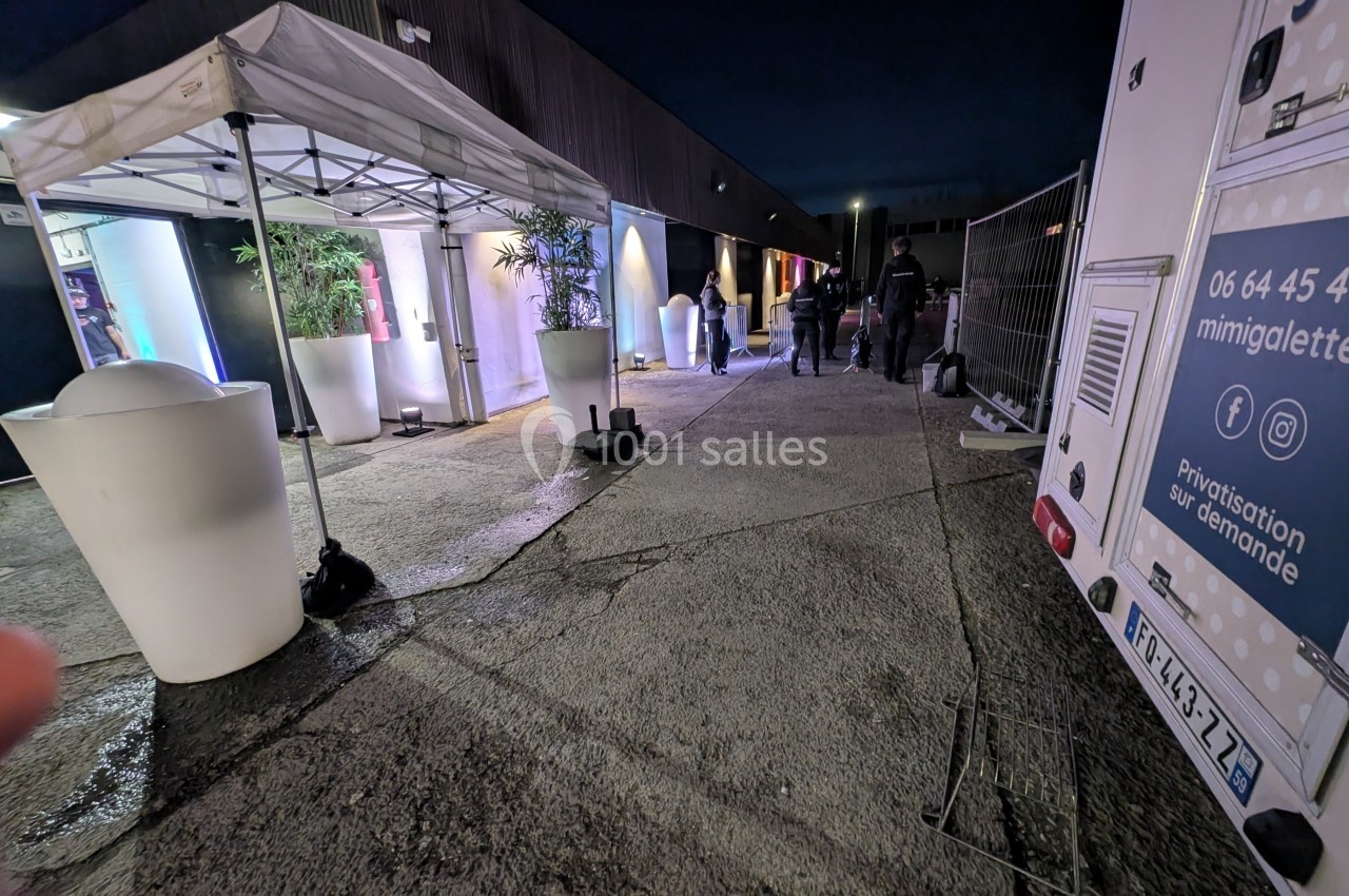 Entrée d'un bâtiment éclairé la nuit avec des tentes blanches, des plantes et des personnes marchant.