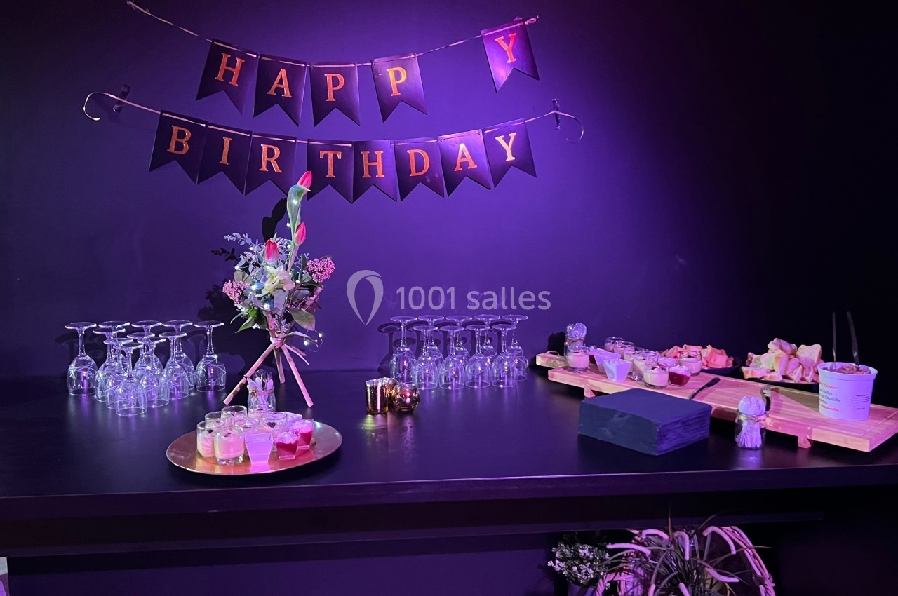 Table décorée pour un anniversaire avec guirlande ’Happy Birthday’, verres, fleurs et plateaux de nourriture.