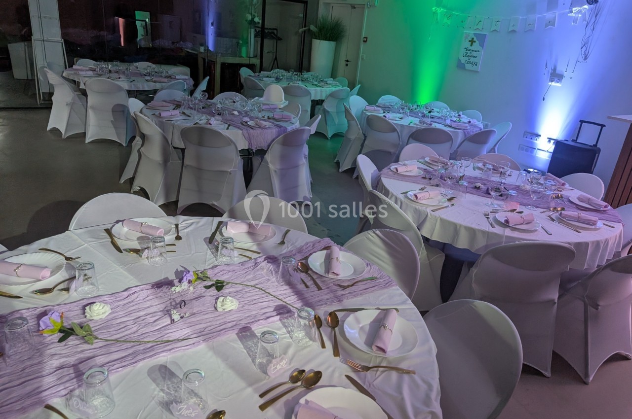 Salle décorée pour un événement avec des tables rondes couvertes de nappes blanches et accents violets.