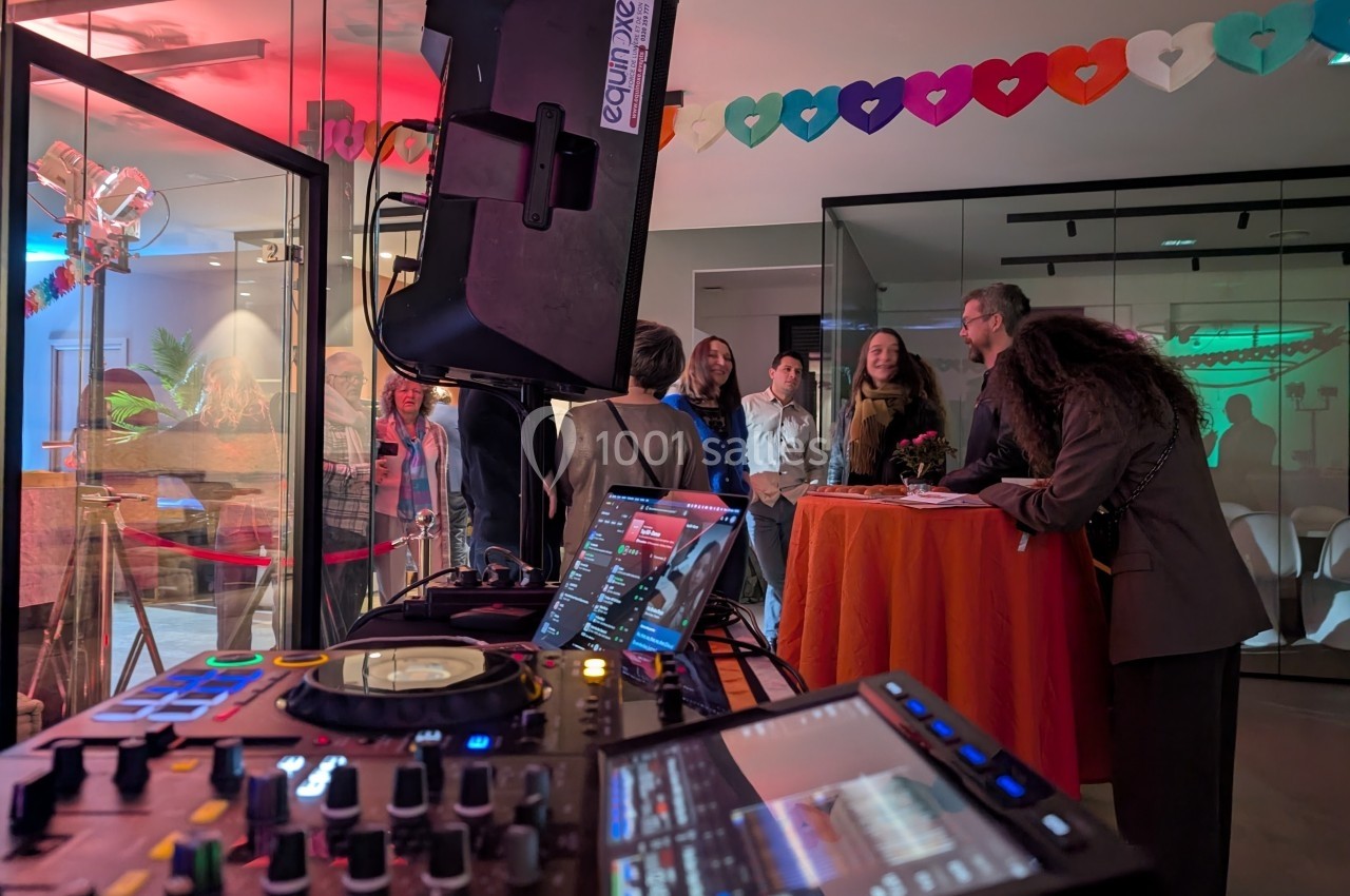Vue d'un équipement de DJ au premier plan avec des invités discutant dans une salle décorée de guirlandes colorées.