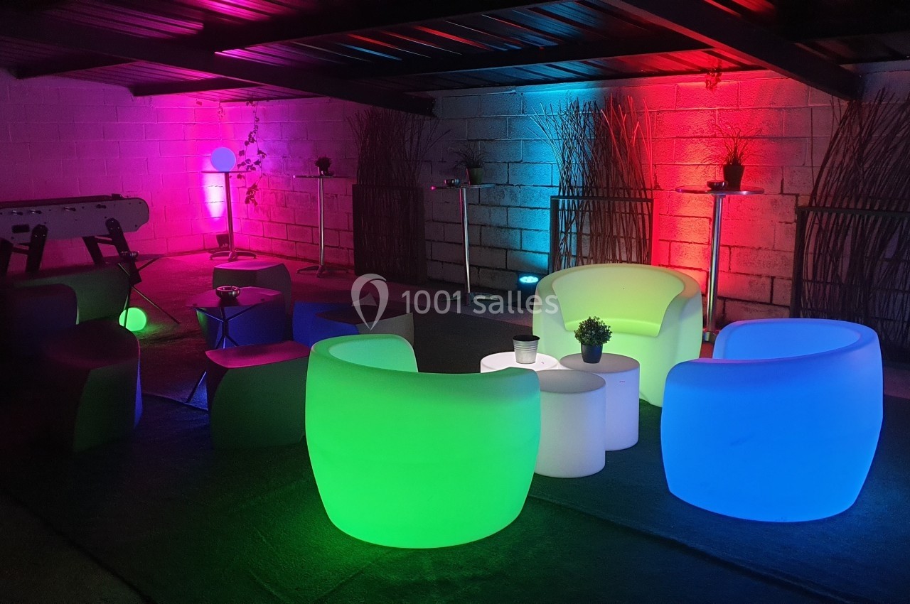 Salle avec des fauteuils lumineux colorés, tables basses blanches et éclairage d'ambiance dans un espace couvert.
