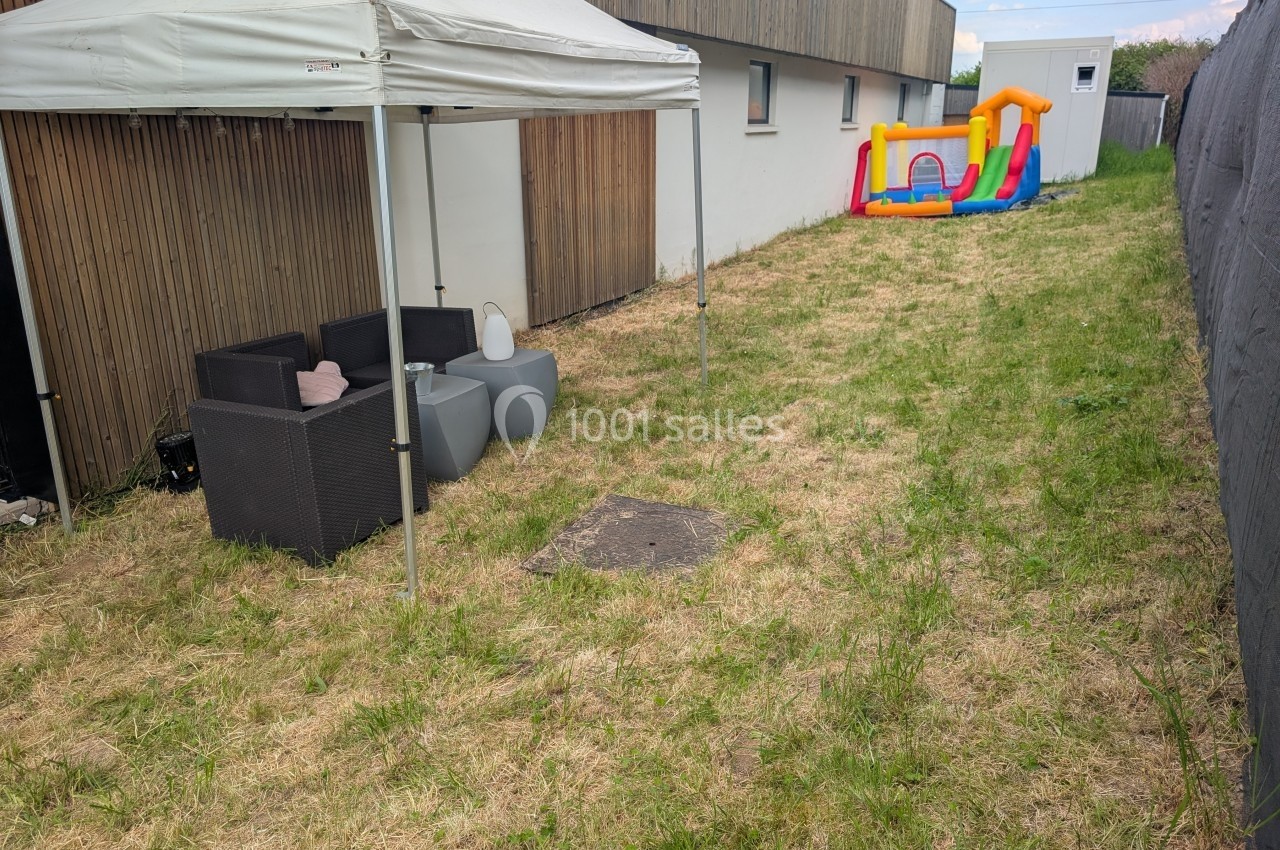 Jardin avec pelouse, tonnelle abritant des fauteuils, petite table et château gonflable coloré en arrière-plan.