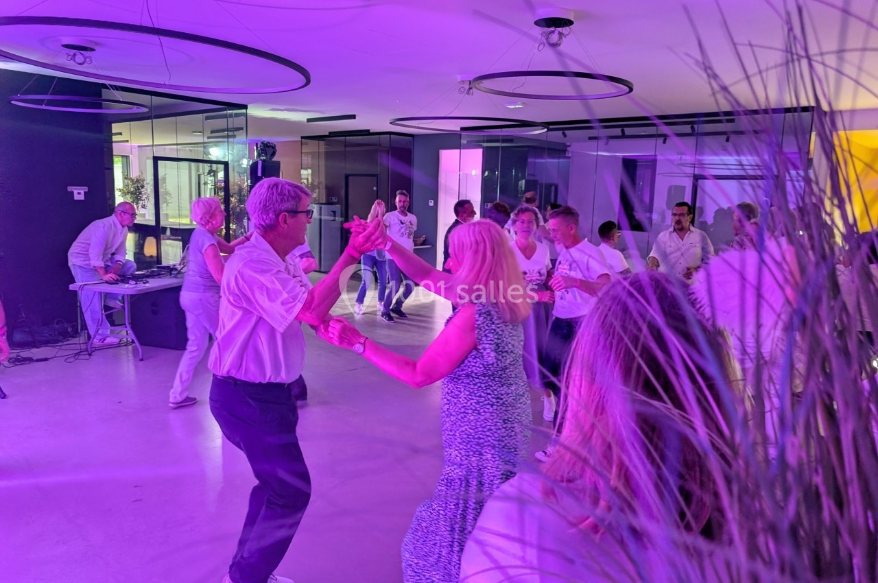 Des personnes dansent en couple dans une salle éclairée par des lumières violettes, avec un DJ en arrière-plan.