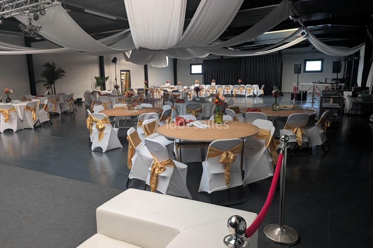 Salle de réception décorée avec des tables rondes, nappes blanches, nœuds dorés et drapés suspendus au plafond.