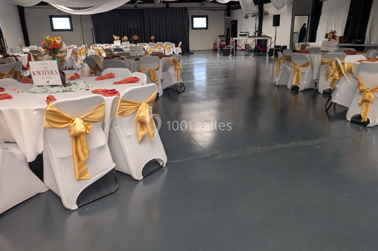 Salle de réception décorée avec des nappes blanches, des chaises ornées de nœuds dorés et des tables dressées.