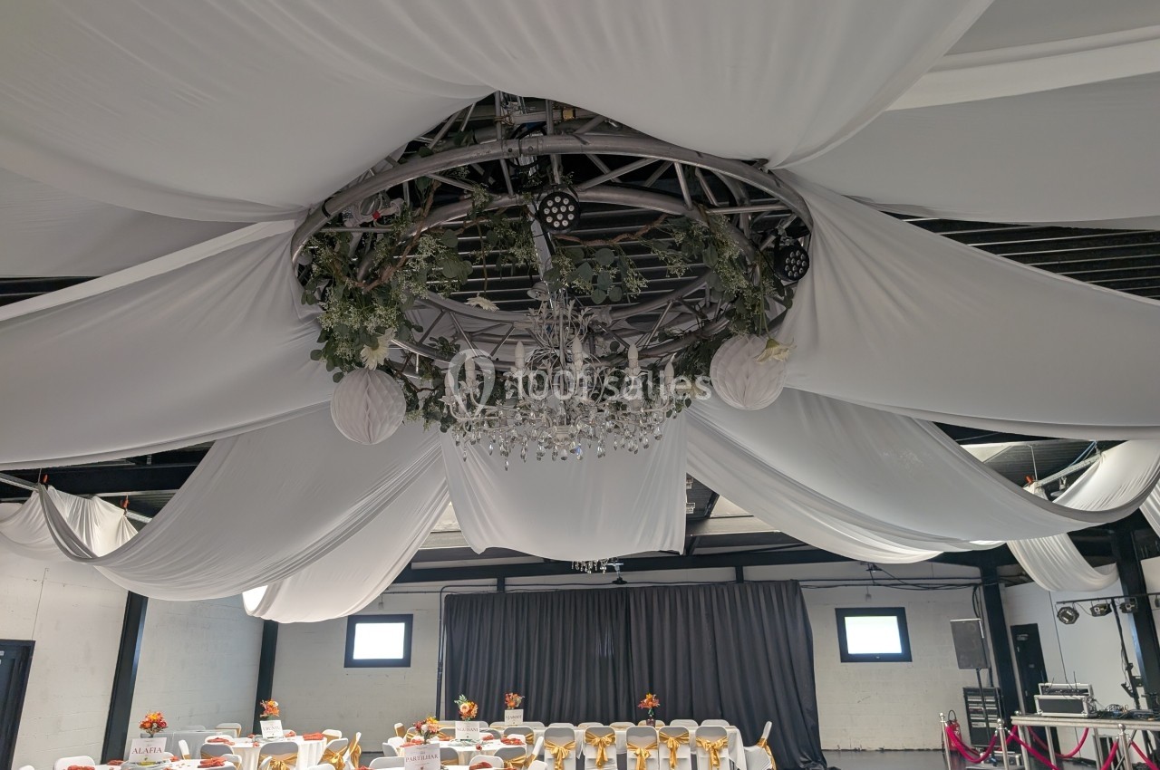 Salle de réception décorée avec des drapés blancs au plafond, un lustre central et des tables dressées.