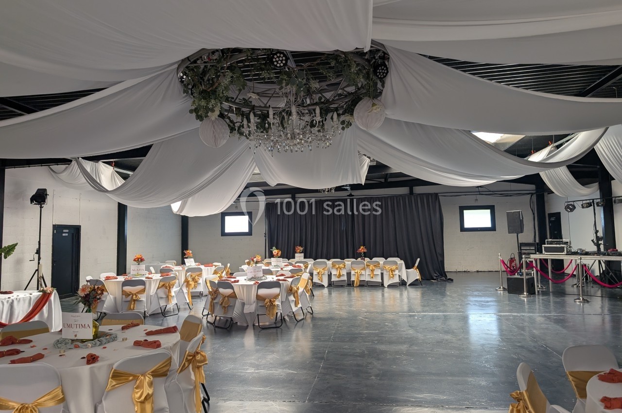 Salle de réception décorée avec des nappes blanches, des chaises ornées de nœuds dorés et un lustre central suspendu.