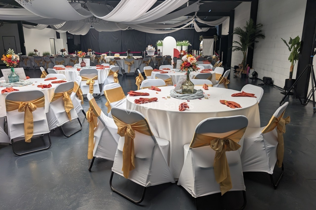 Salle de réception décorée avec des tables rondes, nappes blanches, chaises ornées de rubans dorés et centres de table…