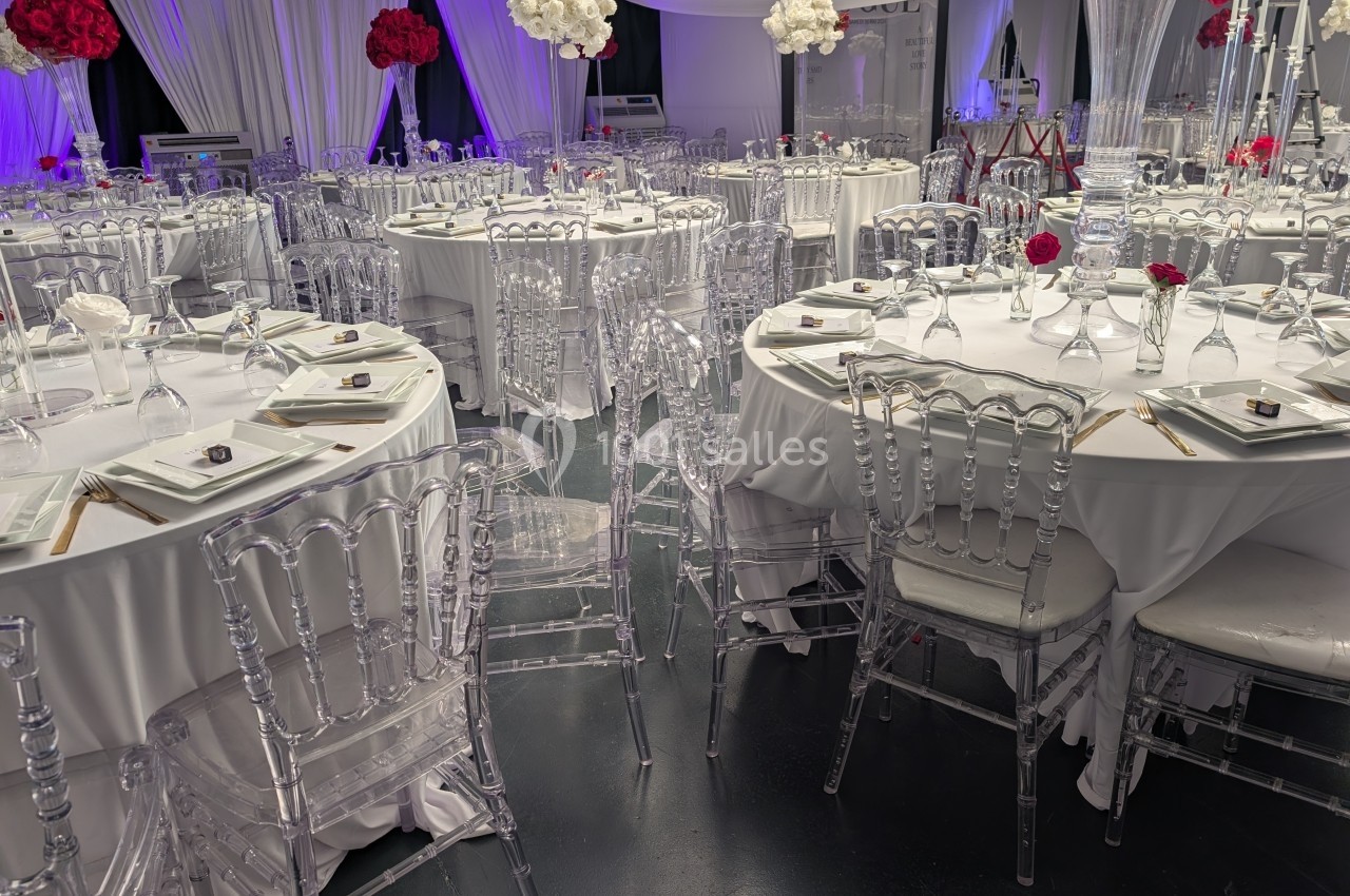Salle de réception décorée avec des tables rondes, nappes blanches, chaises transparentes et centres de table floraux rouges…