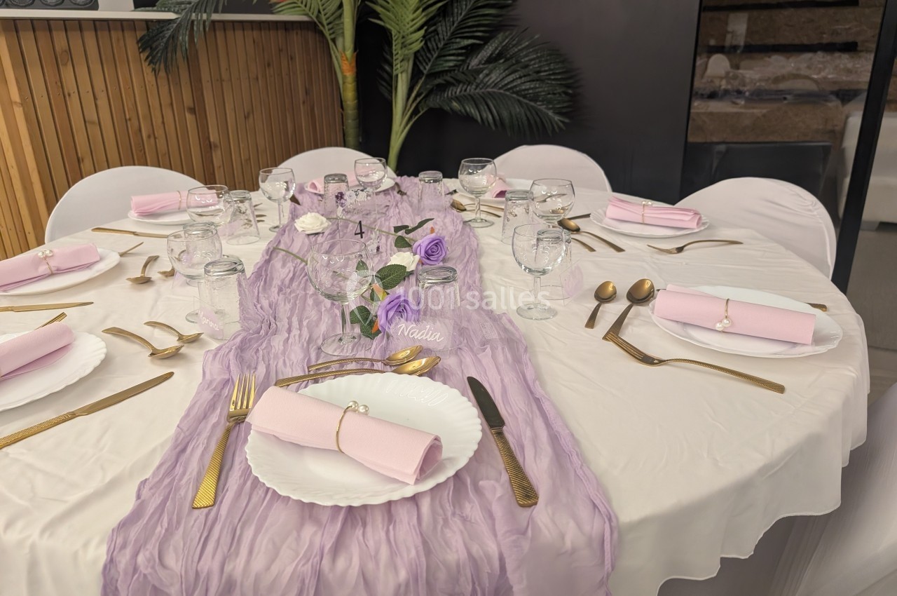 Table décorée avec une nappe blanche, un chemin de table violet, des couverts dorés et des serviettes roses pliées.