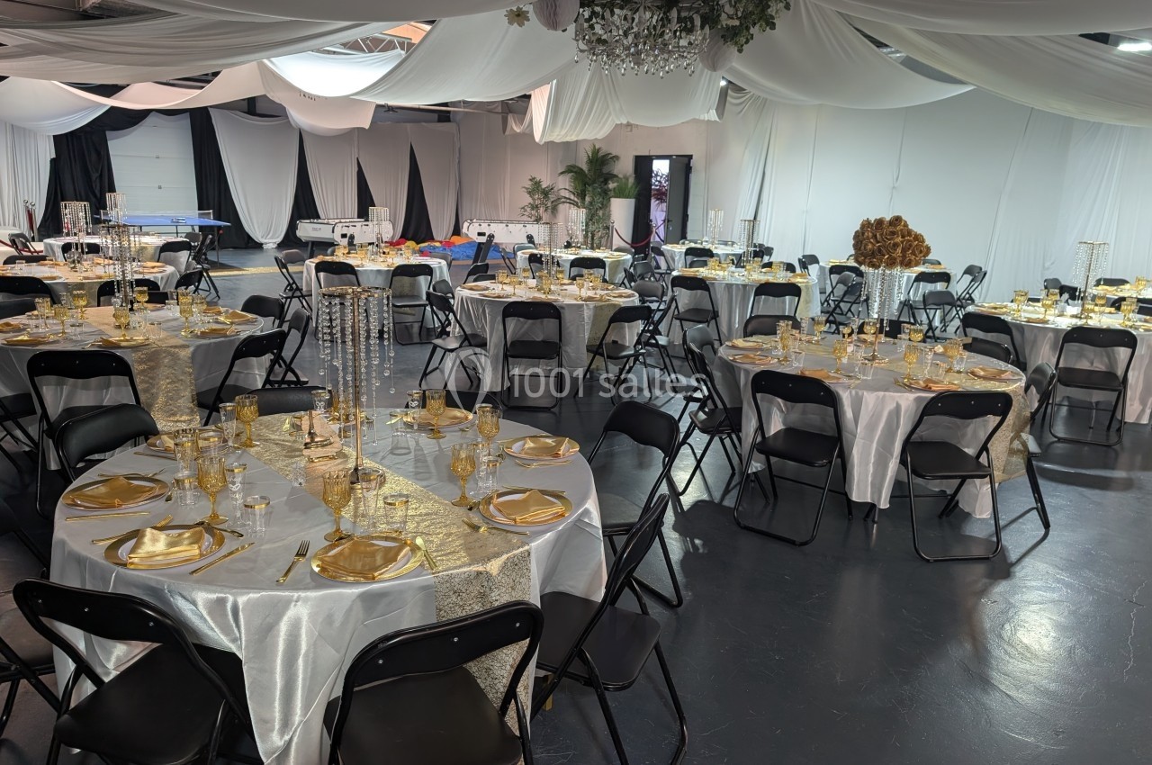 Salle de réception décorée avec des tables rondes dressées de nappes blanches, vaisselle dorée et chaises noires.