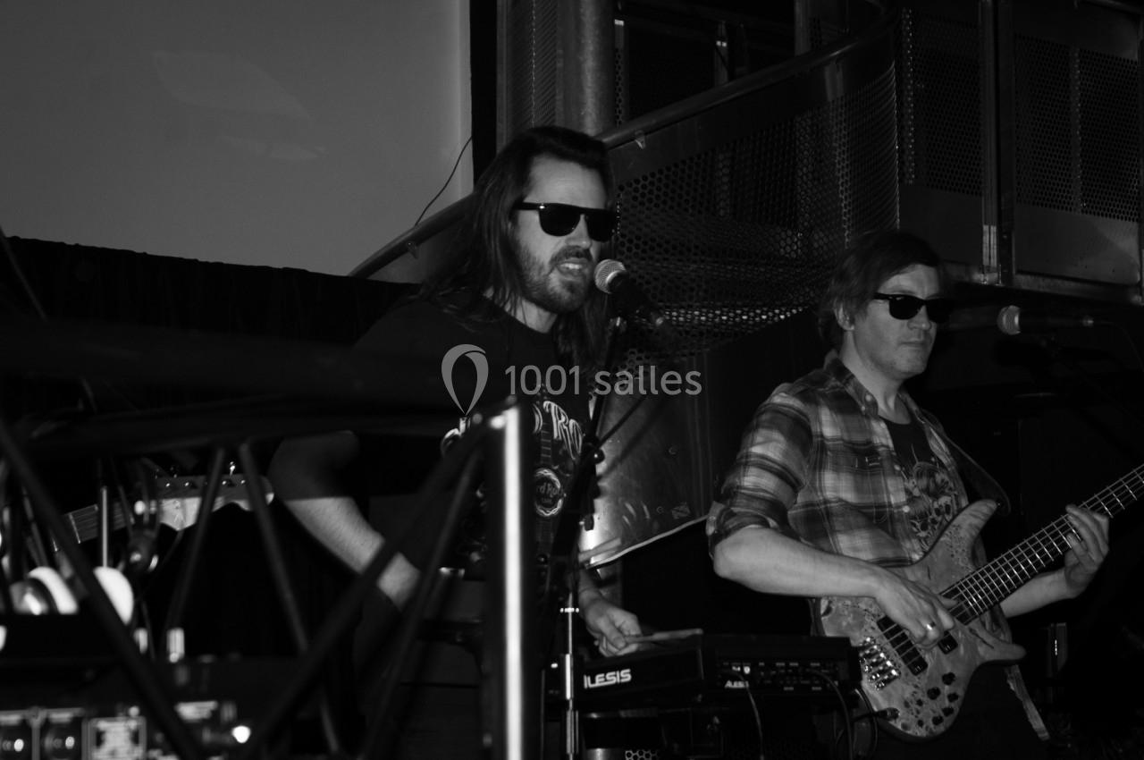 Deux musiciens sur scène, l'un chantant au micro et l'autre jouant de la basse, dans une ambiance sombre.