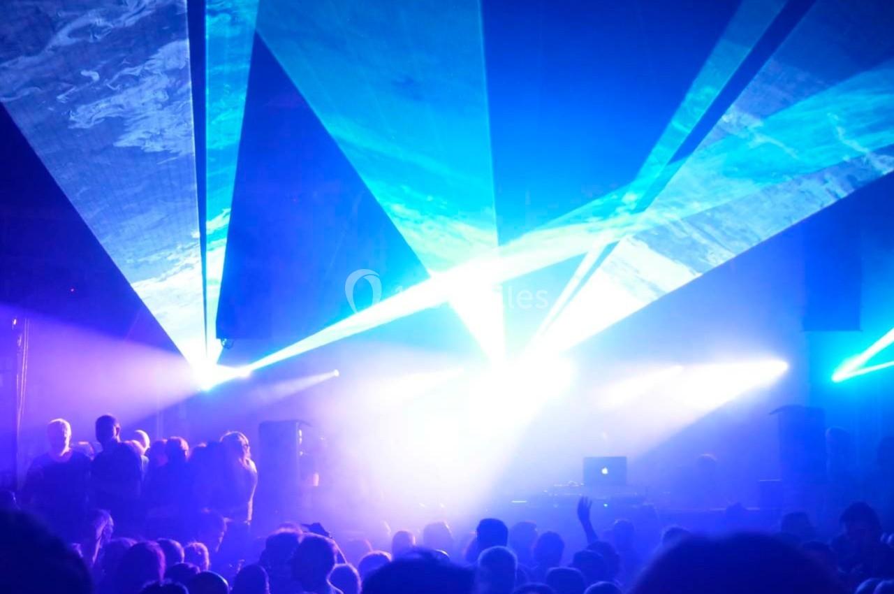 Foule dans une salle sombre illuminée par des faisceaux lumineux bleus et violets, avec un DJ en arrière-plan.