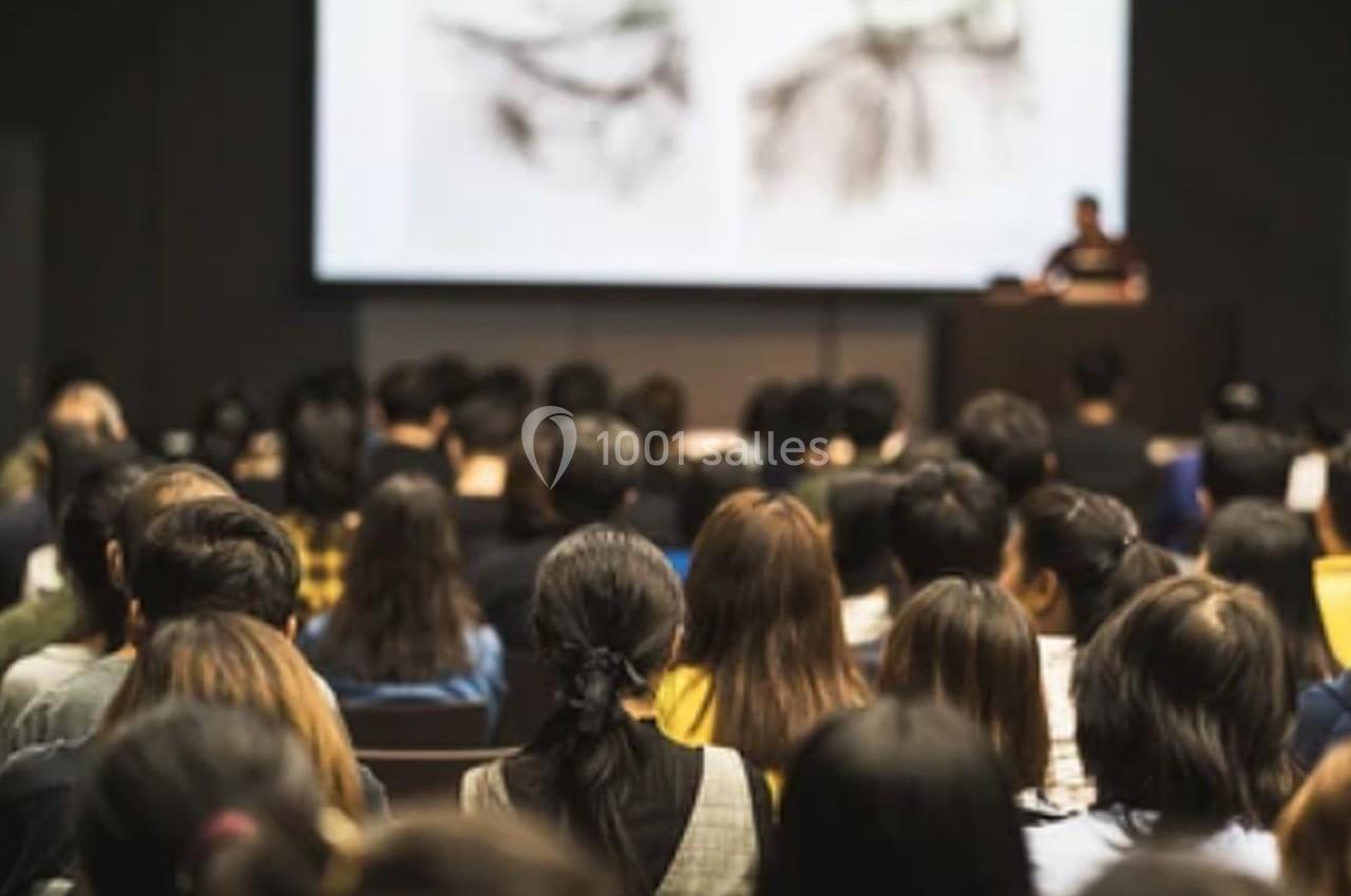Salle comble avec un public attentif face à un intervenant et une présentation projetée sur écran.