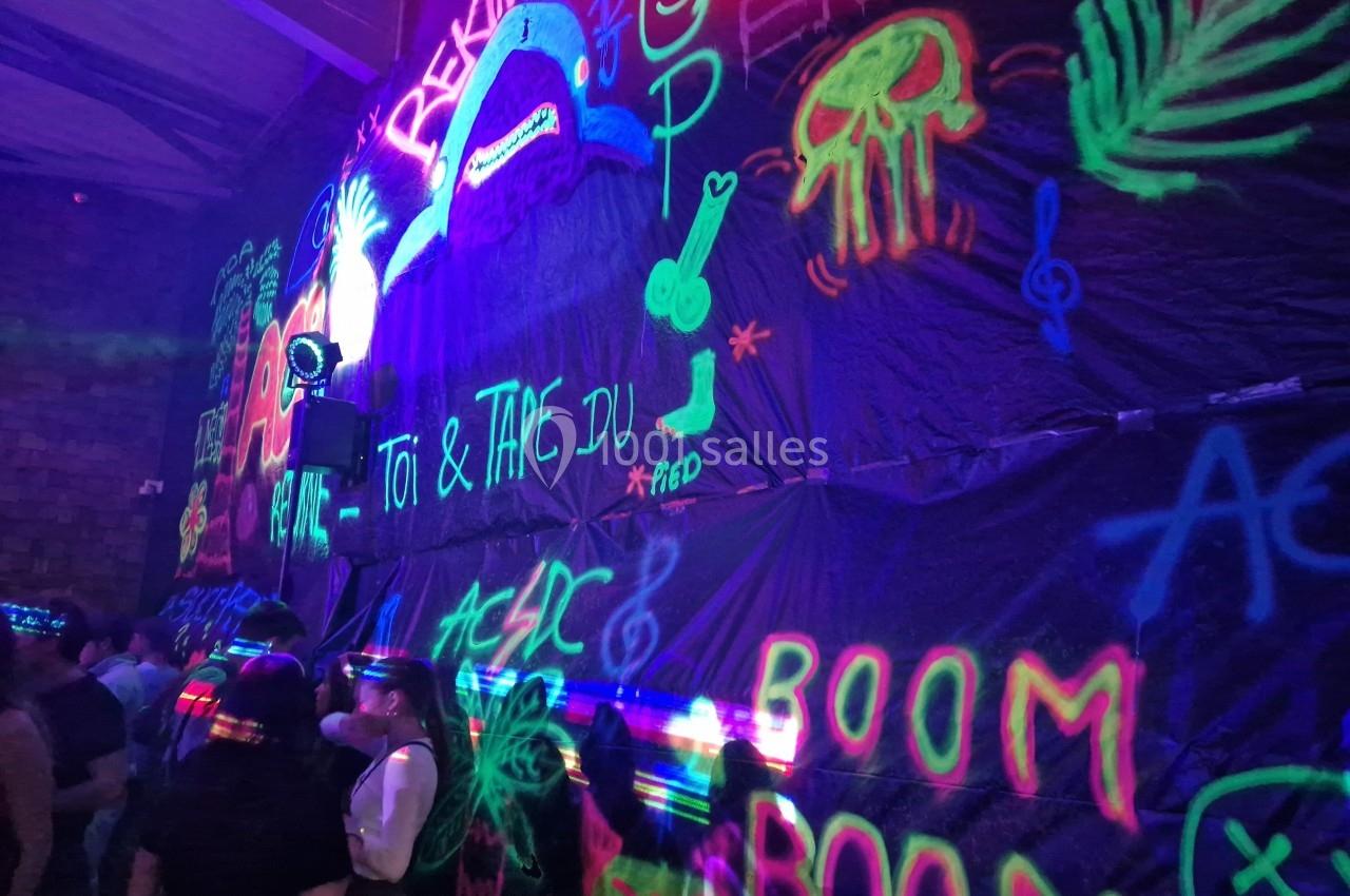 Mur décoré de graffitis fluorescents sous lumière noire, avec des personnes rassemblées au premier plan.