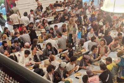 Un grand groupe de jeunes assis dehors sur un sol en béton, près de bâtiments sous un ciel nuageux.