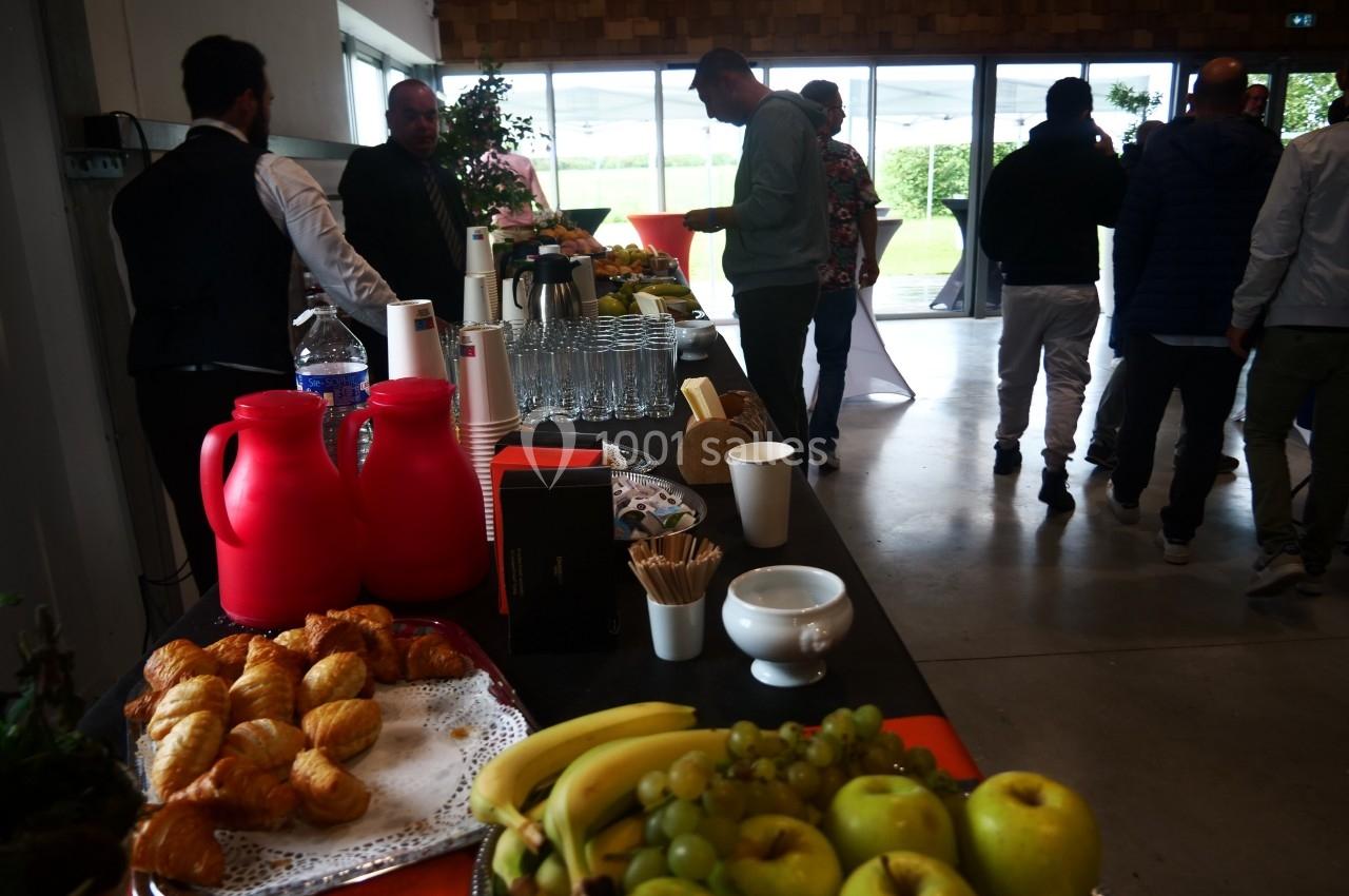 Table de buffet avec viennoiseries, fruits, boissons et convives se servant dans une salle lumineuse.