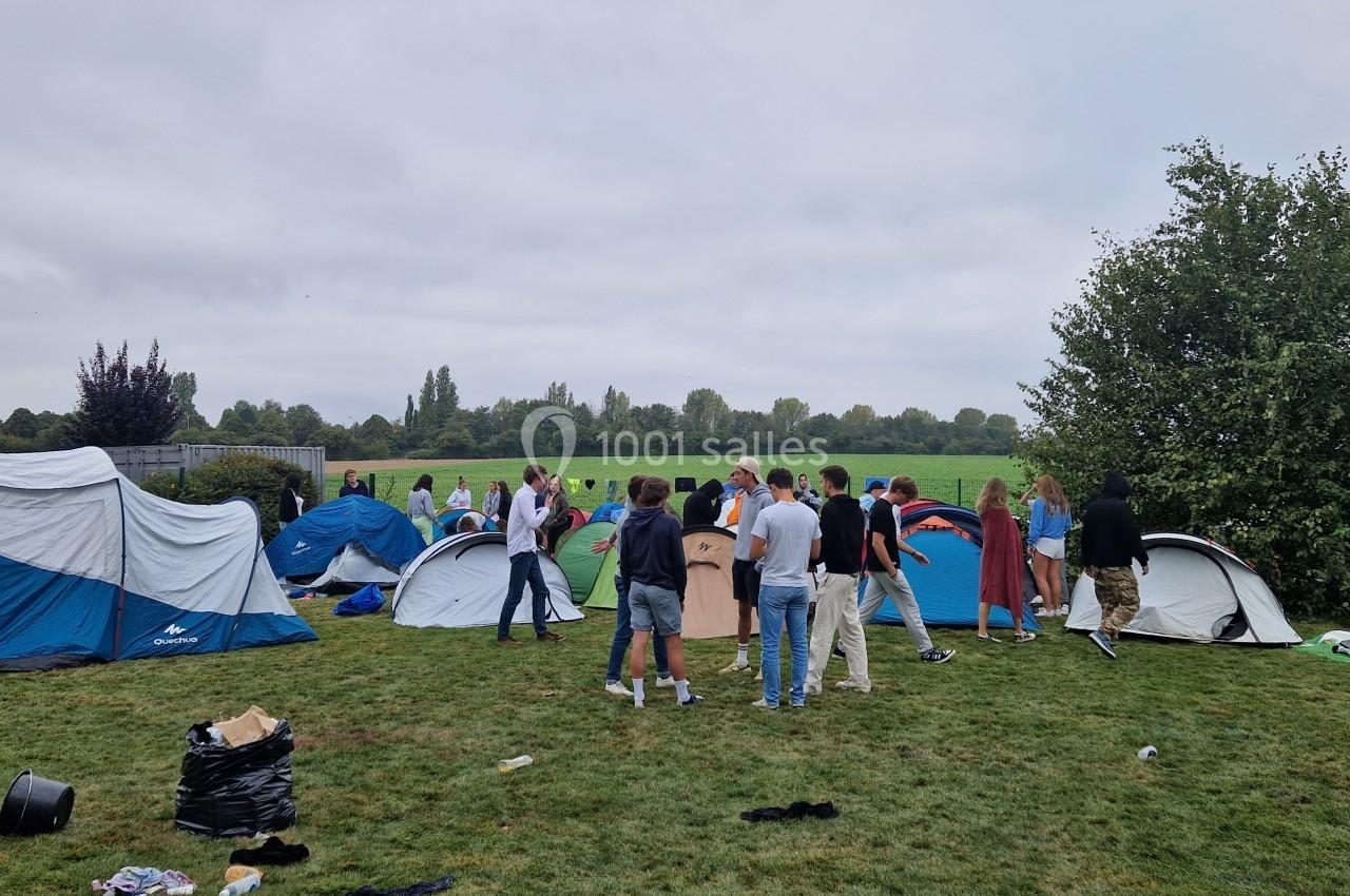 Des personnes montent ou discutent près de tentes sur une pelouse, entourées d'arbres et d'un ciel nuageux.