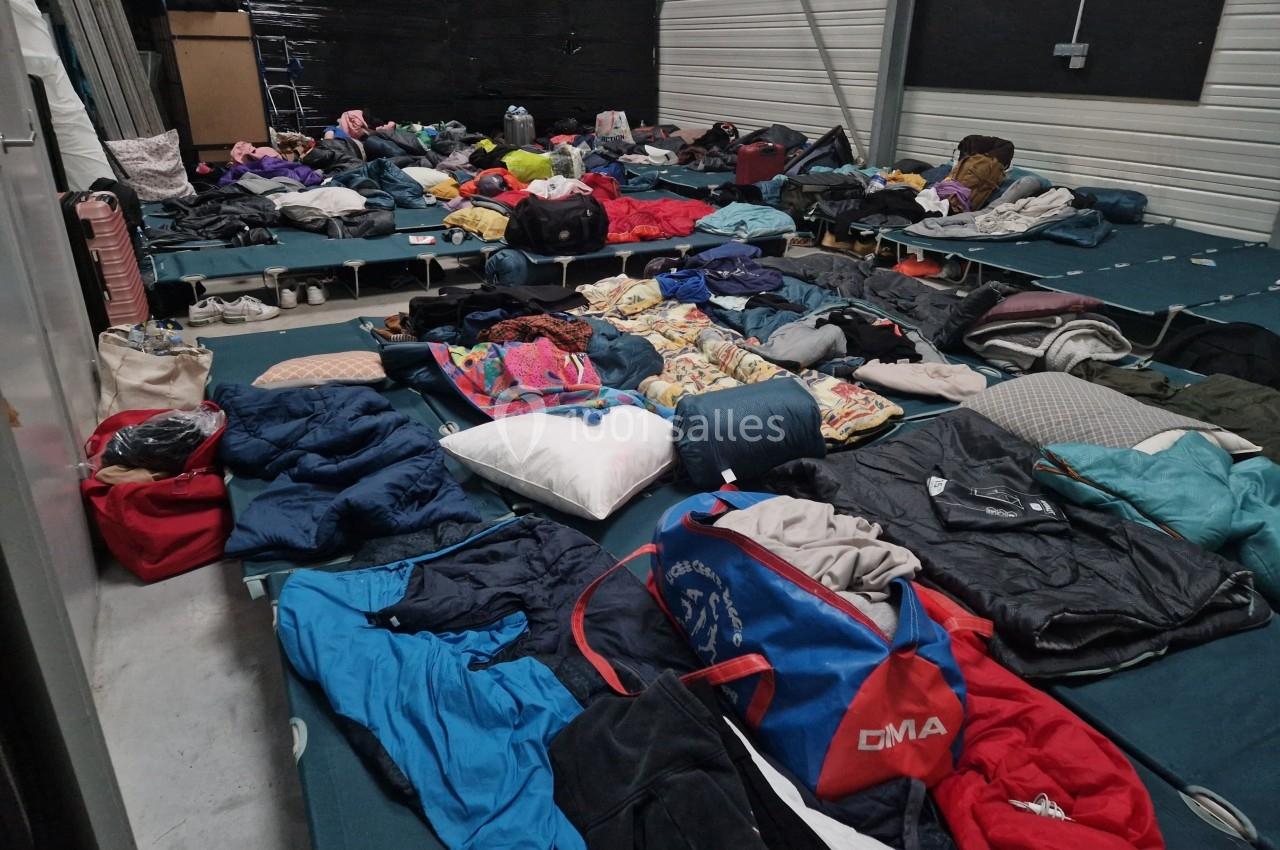 Des matelas au sol recouverts de vêtements, sacs et effets personnels dans un espace intérieur.