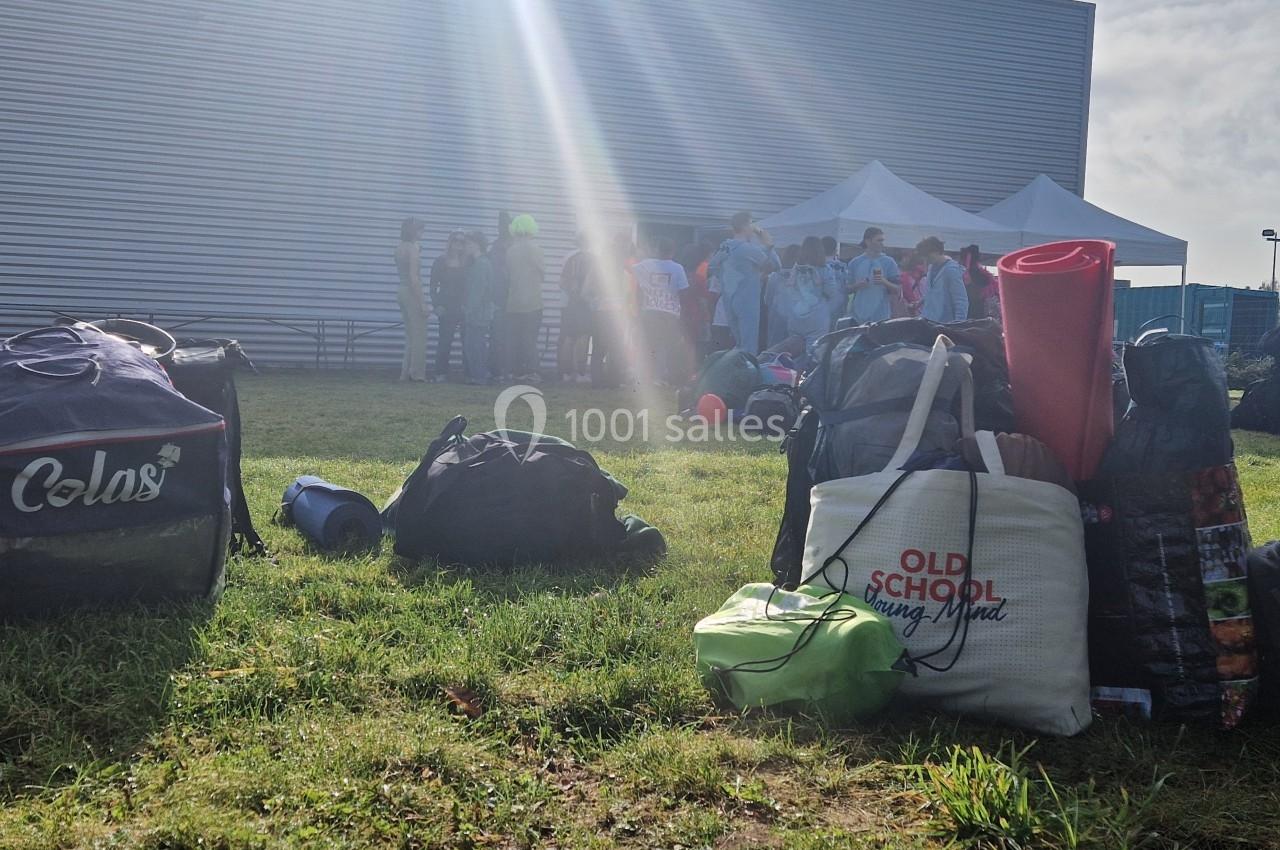 Des sacs et du matériel posés sur une pelouse, avec un groupe de personnes rassemblées près d'une tente blanche.