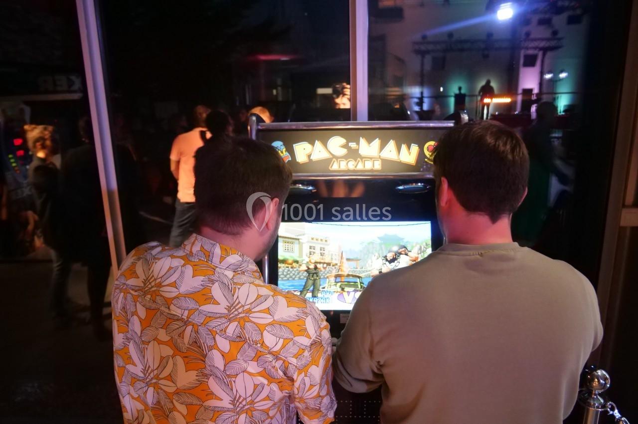 Deux personnes jouent à une borne d'arcade Pac-Man dans un espace intérieur faiblement éclairé.