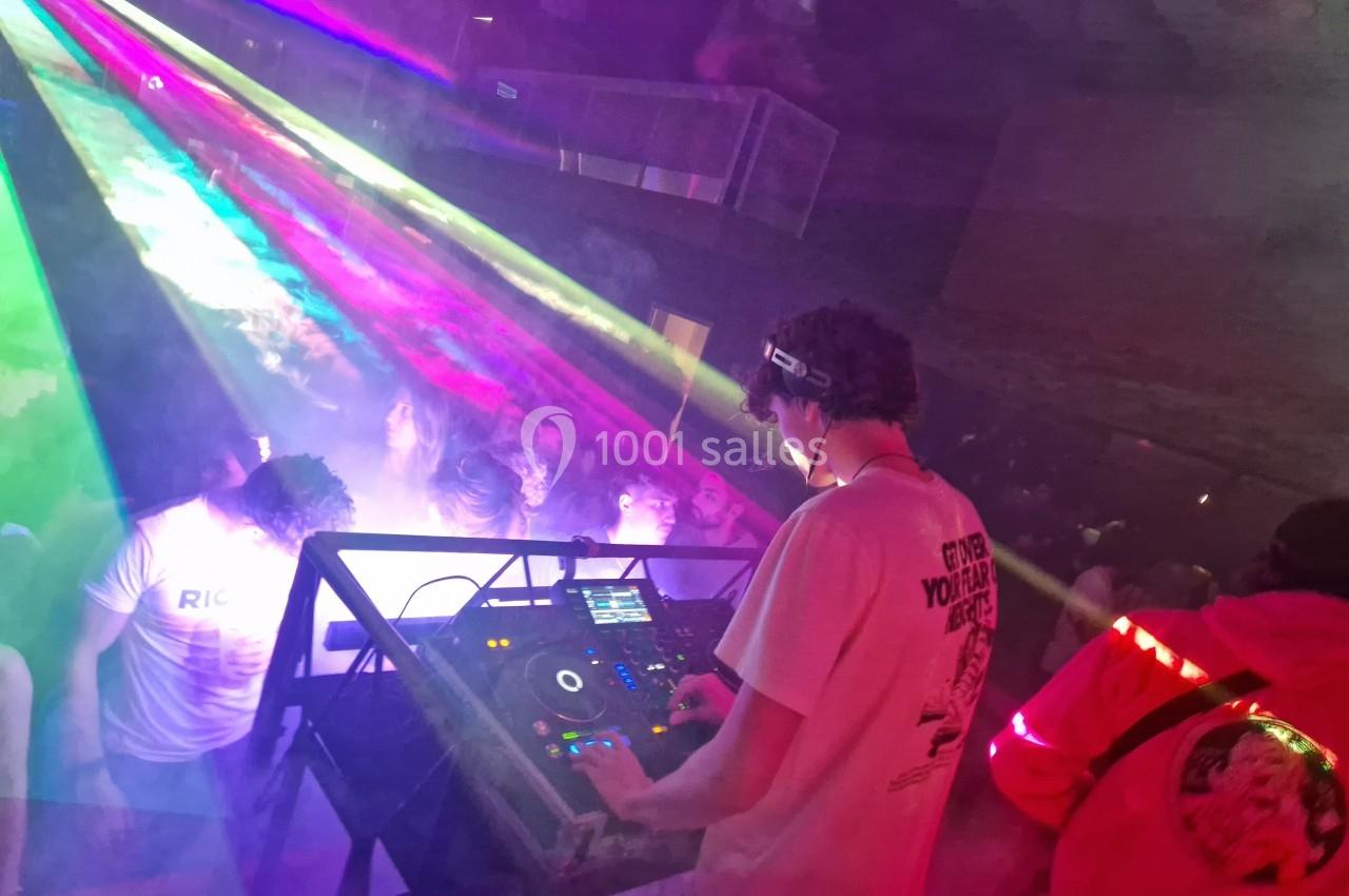 Un DJ mixe sur une scène éclairée par des faisceaux lumineux colorés, face à une foule dans une ambiance festive.