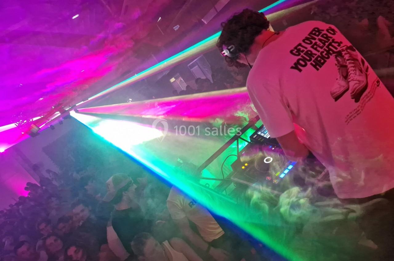 Un DJ mixe sur scène dans une ambiance festive avec des jeux de lumière colorés et une foule rassemblée.