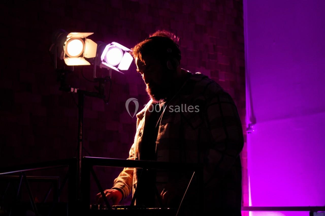 Un DJ en chemise à carreaux mixe sur une table de son, éclairé par des projecteurs dans une ambiance violette.