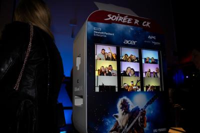 Un grand groupe de jeunes assis dehors sur un sol en béton, près de bâtiments sous un ciel nuageux.