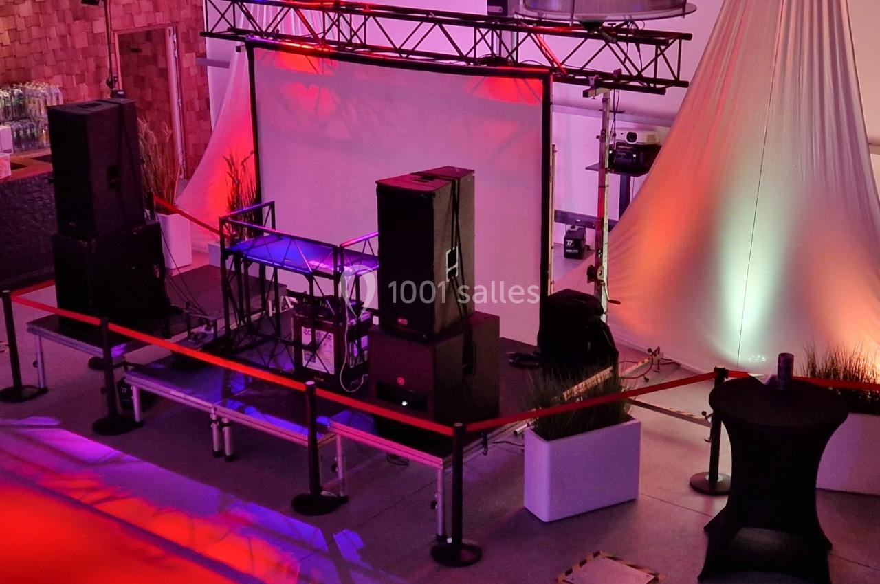 Installation de scène avec projecteurs colorés, écran blanc, enceintes et barrière de sécurité dans une salle événementielle.